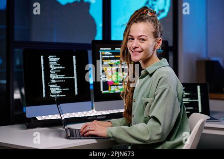 Young successful female programmer looking at camera by workplace with group of desktop computers with coded data on their screens Stock Photo