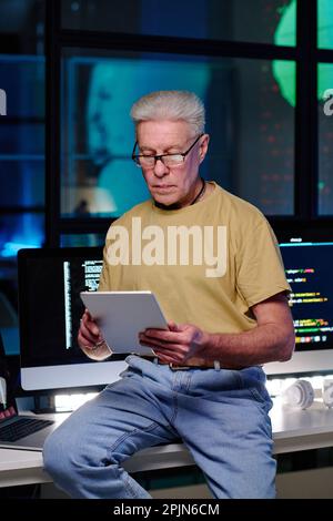 Experienced software developer with tablet sitting on desk against computers with coded data on screens and working late at night Stock Photo