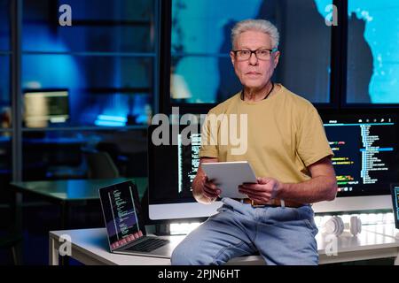 Experienced IT engineer with grey hair using tablet and looking at camera while sitting on desk with computers with coded data on screens Stock Photo