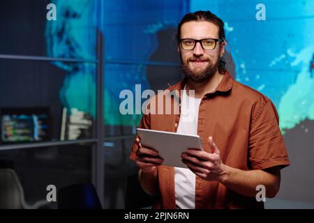 Young smiling IT manager with tablet looking at camera while standing in dark office at night and working over new project or software Stock Photo