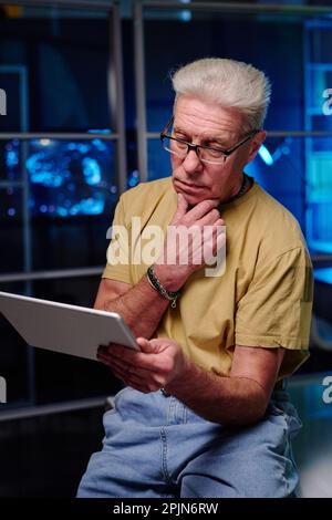 Senior businessman looking through data on tablet screen and thinking of something while standing by workplace in front of camera in office Stock Photo