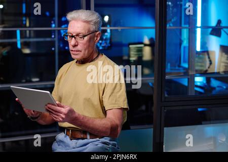 Serious mature programmer in casualwear using tablet while standing in front of camera in dark office at night and developing new software Stock Photo