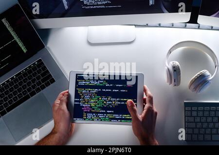 Above view of hands of male IT engineer holding tablet with coded data on screen while sitting by desk with laptop, headphones and keyboard Stock Photo