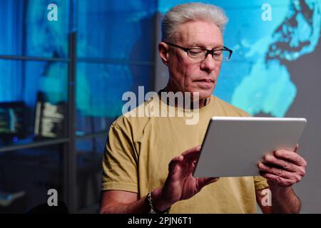 Serious mature businessman with grey hair using digital tablet while standing in frnt of camera in dark office and preparing presentation Stock Photo