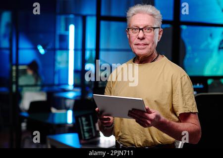 Senior male programmer with tablet standing in front of camera in dark office and looking at you while developing new software Stock Photo