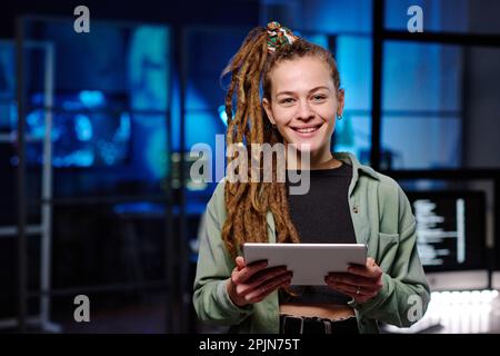 Young cheerful female IT manager with tablet looking at camera while standing in dark office at night and developing new software Stock Photo