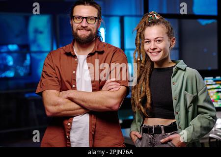 Happy young male and female IT engineers standing in front of camera and looking at you with smiles while working late in dark office Stock Photo