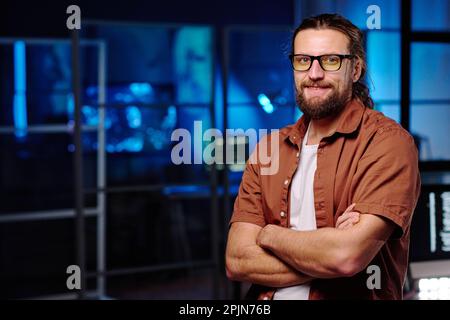 Young smiling male It support manager in eyeglasses crossing arms by chest and looking at camera while standing in dark office Stock Photo