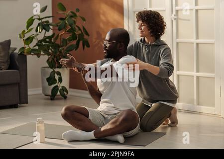 Young woman helping her husband with exercise for stretching arm muscles while sitting behind him on the floor of living room Stock Photo