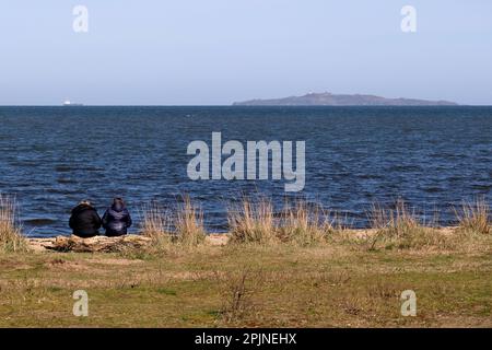 Edinburgh, Scotland, UK. 3rd April 2023. Springtime sunshine attracts visitors to Cramond Foreshore, enjoying the sun and engaging in outdoor activities. Sitting on the beach with a View towards Inchkeith Island, and a container ship heading out into the estuary.. Credit: Craig Brown/Alamy Live News Stock Photo