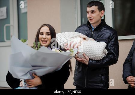 A man holds a baby wrapped in a blanket next to a beautiful women who holds flowers Stock Photo