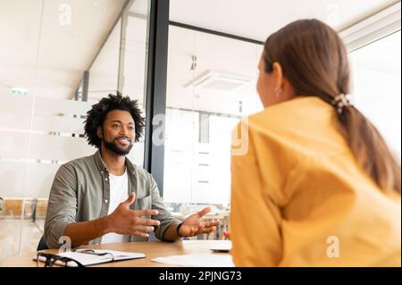 Two happy diverse colleagues African American man and Caucasian woman looking at each other and sitting in office lobby hall, man is gesturing hands Stock Photo