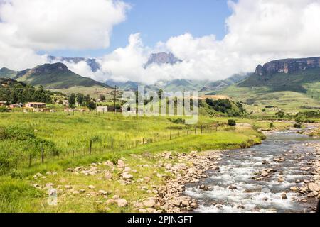 The Tugela river, flowing through a small rural village, with the majestic cliffs of the Drakensberg mountains shrouded in clouds in the Background Stock Photo