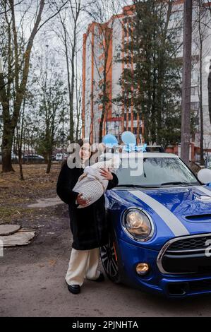 A beautiful woman stands near a car and holds a baby wrapped in a blanket Stock Photo