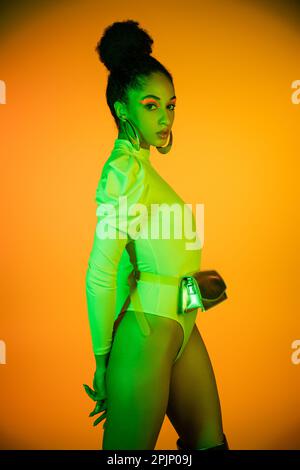 Stylish african american model with neon visage and bodysuit standing on orange background,stock image Stock Photo