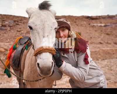 Young woman in warm jacket, posing next to small donkey, leaning on his head. Desert landscape of Petra, Jordan behind her Stock Photo