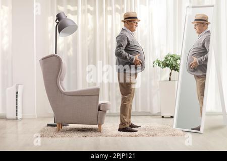 Elderly man with a big belly standing in front of an armchair and looking at a mirror at home Stock Photo