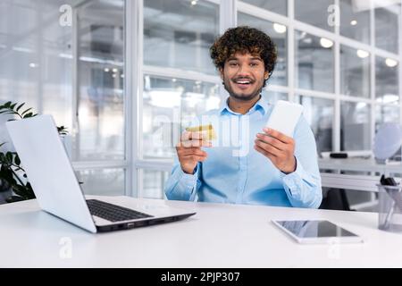 Happy young man sitting in office at desk with phone and credit card. Makes online purchases, pays bills. Smiling looking at the camera. Stock Photo