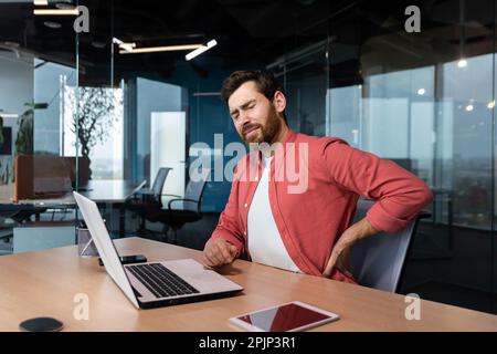Back pain at the workplace. A young male programmer, freelancer sits in the office at the table and holds his back twisted, stretched, tension pain. Tired from an overtime day. Stock Photo