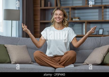 Senior beautiful woman sitting on sofa at home and doing yoga, online, remotely. Talk about mental and psychological health. She closed her eyes in the lotus position, relaxed. Stock Photo