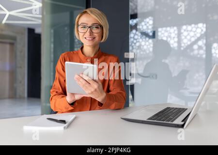 Portrait of a young female student sitting in an office, co-working space, library at a table with a laptop and using a tablet to study. Smiling, she looks at the camera. Stock Photo