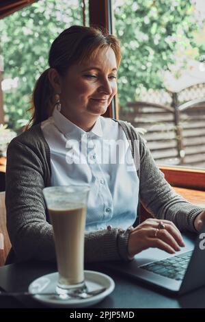 Smiling happy businesswoman working remotely on her laptop computer managing her work typing on laptop keyboard sitting at the table in a cafe Stock Photo