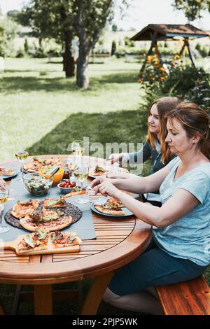 Family and friends having meal - pizza, salads, fruits and drinking white wine during summer picnic outdoor dinner in a home garden. Close up of peopl Stock Photo