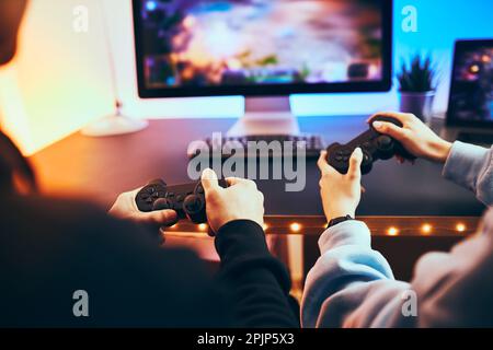 Friends playing video game at home. Gamers holding gamepads sitting at front of screen. Streamers girl and boy playing online in dark room lit by neon Stock Photo