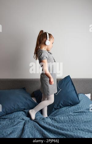 Little girl singing holding headphones cord imitating herself a real singer. Child having fun jumping dancing listening to music on bed in bedroom at Stock Photo