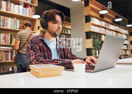 Student learning in university library. Young man preparing for test on laptop. Man listening to online course. Focused student studying for college e Stock Photo