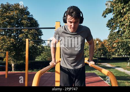 Young man doing dips on parallel bars during his workout in a modern calisthenics street workout park. Man listening to music wearing headphones Stock Photo