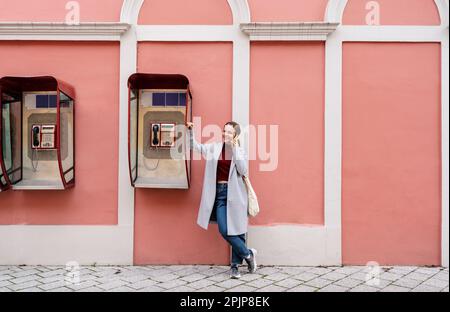 Woman standing next to the street payphone and talking on mobile phone. Stock Photo