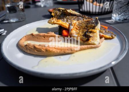 Roasted cod fillet on chestnut puree and creamy sauce on white plate, glass of water, outdoor restaurant. Stock Photo