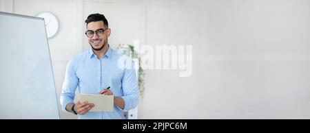 Cheerful confident arab millennial guy teacher in glasses stands near whiteboard in office class interior, panorama Stock Photo