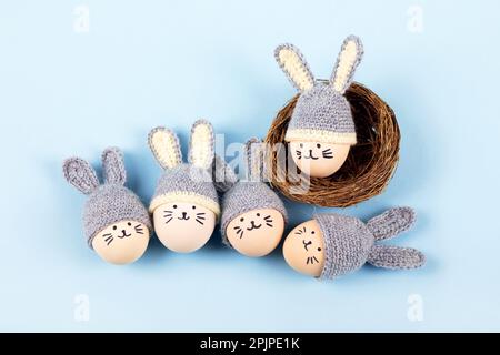 Five Easter eggs with funny faces and gray crocheted hats with bunny ears in a decorative nest of sisal on a blue background. Happy Easter concept. Gr Stock Photo