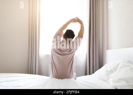 Back view of man wearing pajamas sitting on bed, stretching Stock Photo