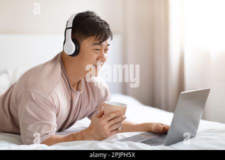 Positive asian man using laptop and headset in bed Stock Photo