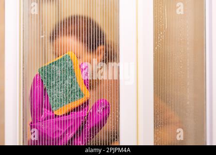 A woman's hand in latex gloves holds a sponge and washes the glass walls of the shower cabin in the bathroom. Selective focus, copy space. Stock Photo