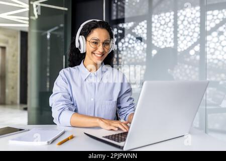 Young beautiful Indian female programmer working inside office with laptop, woman in headphones listening to audio podcast and online course, typing on keyboard and reaching out, female engineer . Stock Photo