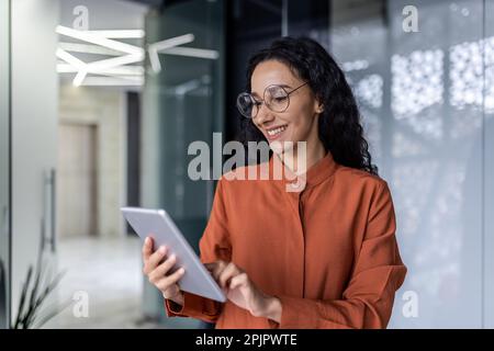 Latin american female programmer inside office standing near window using tablet computer for software testing and developing applications online. Stock Photo