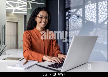 Beautiful and successful hispanic business woman inside office working with laptop, office worker with curly hair smiling woman boss happy with achievement results at workplace. Stock Photo