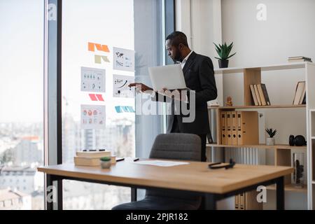 Multiethnic professional in formal attire handling laptop with one hand while studying graphs and charts on glass panel. Efficient systems analyst managing new project using data from infographics. Stock Photo