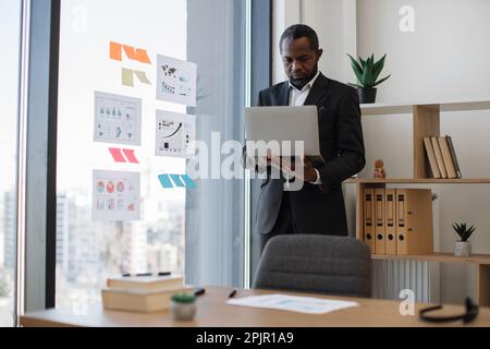 African american businessman typing on computer keyboard while standing near glass window with visual representation of data. Proficient manager trying to gain better insight about project at work. Stock Photo