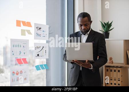 African american businessman typing on computer keyboard while standing near glass window with visual representation of data. Proficient manager trying to gain better insight about project at work. Stock Photo
