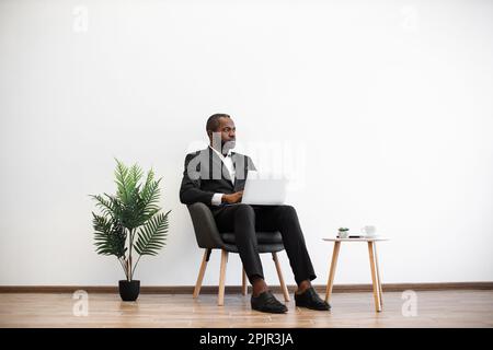 Focused african american man sitting at office and working on modern laptop. Handsome young male wearing black formal suit browsing internet indoors. Concept of people and technology. Stock Photo