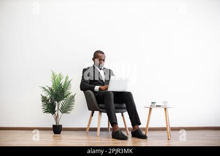 Focused african american man sitting at office and working on modern laptop. Handsome young male wearing black formal suit browsing internet indoors. Concept of people and technology. Stock Photo