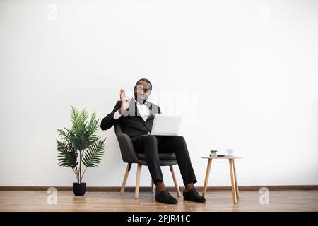 Confused african man in black suit having video conference on wireless laptop while working at office. Intense conversation with coworkers on distance. Modern lifestyles of business people. Stock Photo