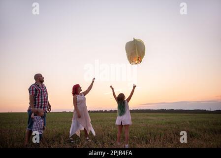 Diversity family launch yellow sky lantern in field at sunset. Mother, father holding Chinese paper lantern, daughter, son wave hands. Making wishes f Stock Photo