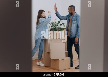 A young couple high-fives as they stand in the living room, the hallway of their home, next to cardboard boxes of belongings. Stock Photo