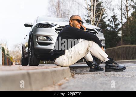 Stylish young adult man in sunglasses sitting on pavement in front of his expensive car and having a phone call. Outdoor full-length shot. High quality photo Stock Photo
